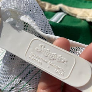 May include: A white plastic adjustable snapback strap on a hat, featuring the embossed text 'Swingster' above 'Shawnee Mission, Kansas, USA'. The strap is attached to a white mesh panel, with a portion of a green fabric hat visible in the background.
