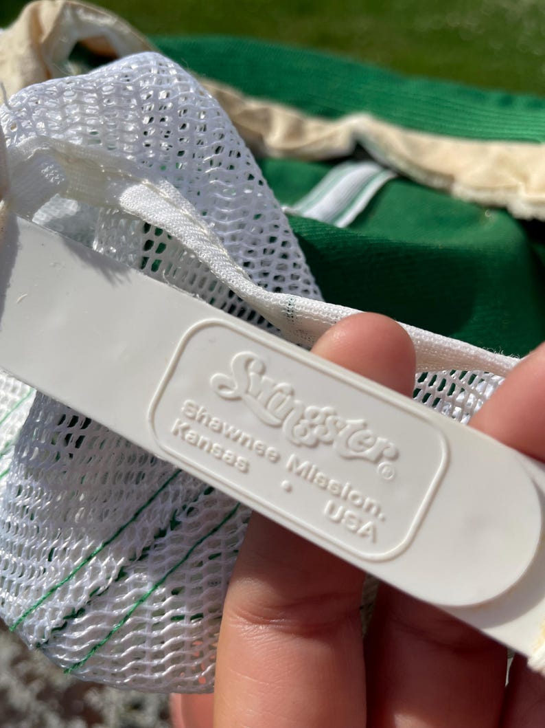 May include: A white plastic adjustable snapback strap on a hat, featuring the embossed text 'Swingster' above 'Shawnee Mission, Kansas, USA'. The strap is attached to a white mesh panel, with a portion of a green fabric hat visible in the background.