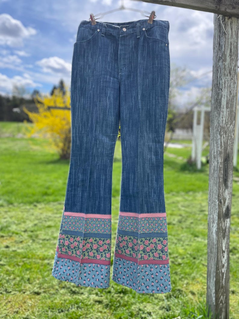Pu&ograve; includere: Jeans blu a zampa con motivo floreale sull'orlo. Il motivo floreale comprende rose rosa, foglie verdi e uno sfondo azzurro. I jeans sono appesi a un filo.
