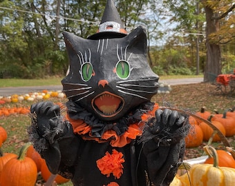 May include: A black cat figurine wearing a pointed black witch hat with a silver band, green eyes, and an open mouth. The cat has a black ruffled collar with orange accents, black sleeves with frayed edges, and two orange pom-poms on the front.