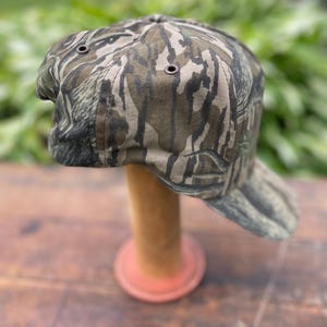 Op de afbeelding: Een baseballpet met een bruin, zwart en beige camouflagepatroon. De pet heeft een gestructureerde kroon met donkergekleurde metalen oogjes voor ventilatie en een gebogen klep. De stof vertoont een gedetailleerde, organische print.