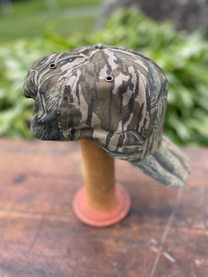 Op de afbeelding: Een baseballpet met een bruin, zwart en beige camouflagepatroon. De pet heeft een gestructureerde kroon met donkergekleurde metalen oogjes voor ventilatie en een gebogen klep. De stof vertoont een gedetailleerde, organische print.