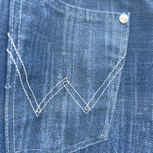 Pu&ograve; includere: Primo piano di una tasca in denim blu con cuciture bianche e un bottone argentato. La tasca presenta un design a "W" cucito in bianco. Il denim ha un aspetto sbiadito.