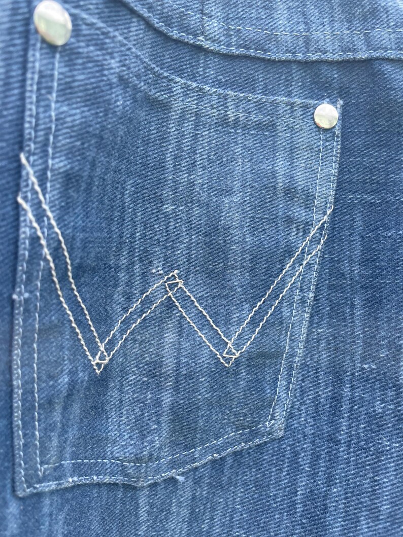Pu&ograve; includere: Primo piano di una tasca in denim blu con cuciture bianche e un bottone argentato. La tasca presenta un design a "W" cucito in bianco. Il denim ha un aspetto sbiadito.