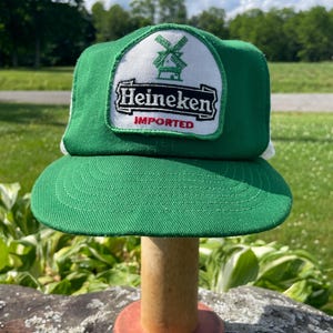 May include: A green and white trucker hat featuring a patch on the front panel. The patch displays a green windmill icon above the black text Heineken and the red text IMPORTED, all set against a white background with a green border.
