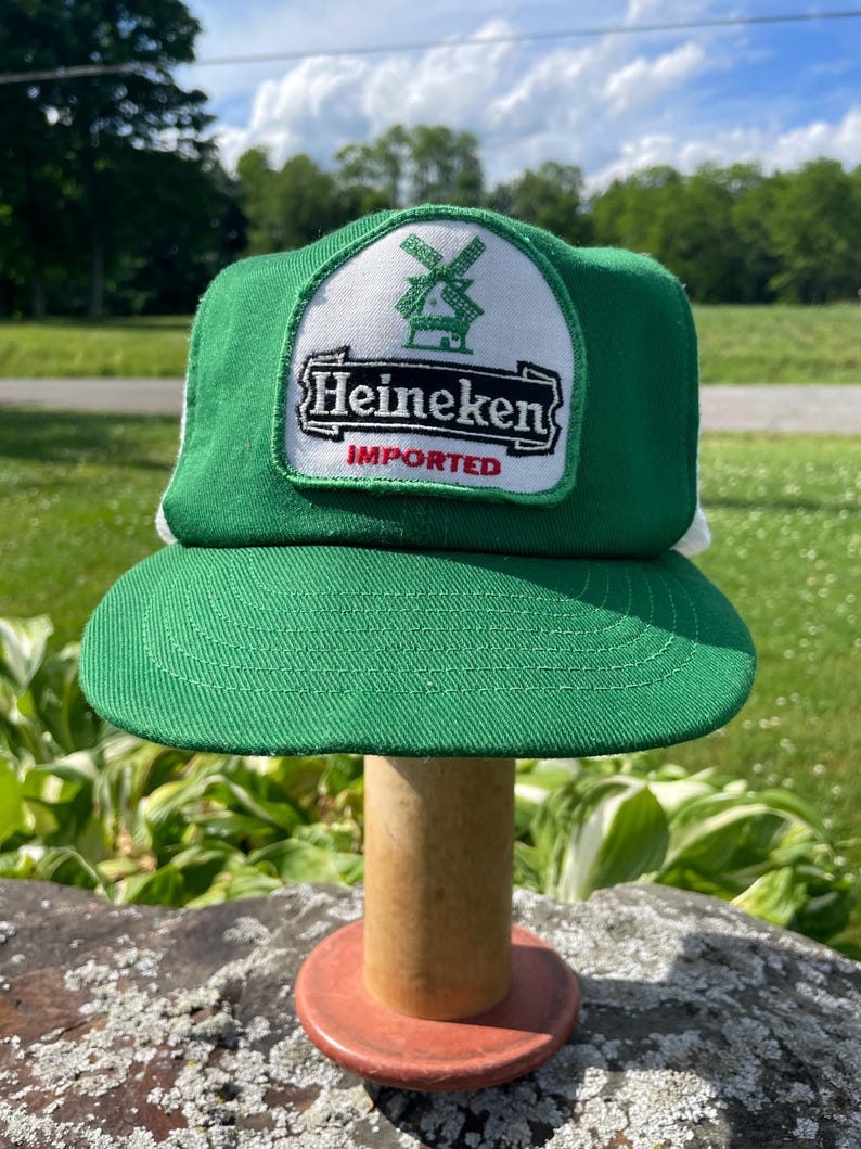 May include: A green and white trucker hat featuring a patch on the front panel. The patch displays a green windmill icon above the black text Heineken and the red text IMPORTED, all set against a white background with a green border.