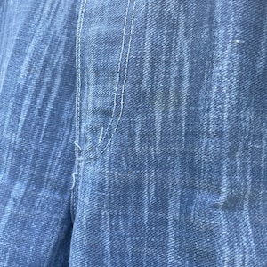 Pu&ograve; includere: Primo piano di tessuto denim blu con trama visibile e cuciture bianche. L'immagine evidenzia i dettagli del materiale denim, mostrando l'intreccio e le variazioni di colore. La cucitura &egrave; centrata e verticale.