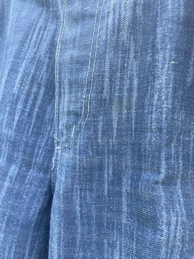 Pu&ograve; includere: Primo piano di tessuto denim blu con trama visibile e cuciture bianche. L'immagine evidenzia i dettagli del materiale denim, mostrando l'intreccio e le variazioni di colore. La cucitura &egrave; centrata e verticale.