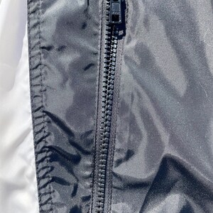 Puede incluir: Primer plano de una chaqueta negra con una cremallera negra. La cremallera est&aacute; centrada y corre verticalmente. La chaqueta tiene una textura brillante y suave. El lado izquierdo de la chaqueta es blanco.