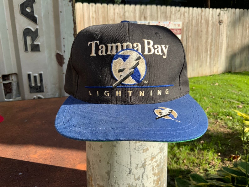 May include: A black and blue baseball cap with the embroidered text "Tampa Bay Lightning" and a lightning bolt logo.