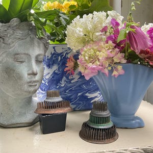 May include: A gray stone-look head planter, a blue and white marbled cylindrical vase, a light blue fan-shaped vase with a floral arrangement, and two metal flower frogs with copper-toned bases and needle-like pins for floral arranging.