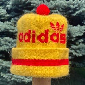May include: A bright yellow knit beanie featuring a red pom-pom on top and a folded cuff. The word adidas is knitted in red across the front, accompanied by the red adidas trefoil logo. A horizontal red stripe encircles the center of the cuff.