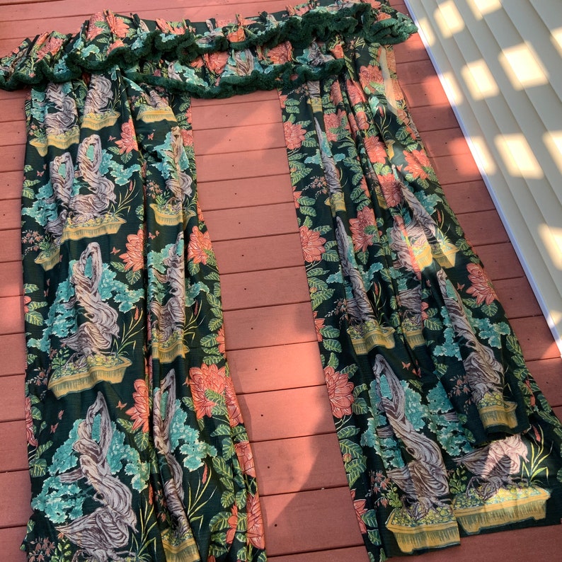May include: A pair of green curtains with a floral pattern featuring pink flowers, green leaves, and brown tree branches. The curtains have a ruffled top edge.