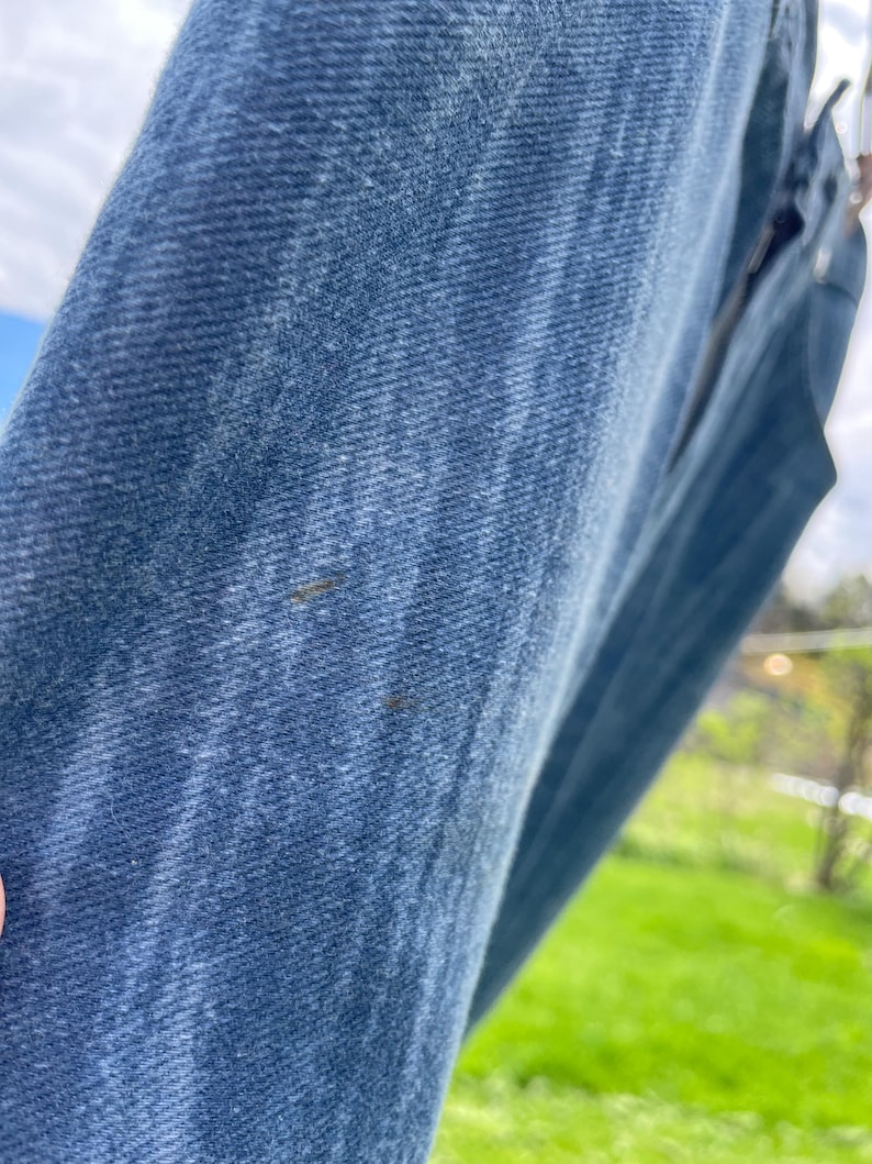 Pu&ograve; includere: Primo piano di jeans di denim blu, che mostra la trama e i dettagli del tessuto. I jeans sono appesi all'esterno, con uno sfondo verde sfocato. Il denim ha un aspetto classico e usurato.