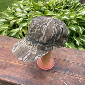 Op de afbeelding: Een baseballpet met een camouflagepatroon in bruin-, beige- en donkergroene tinten. De pet heeft een gestructureerde kroon, een gebogen klep en een klein donker oogje aan de zijkant. De stof is voorzien van een gedetailleerde bosprint.