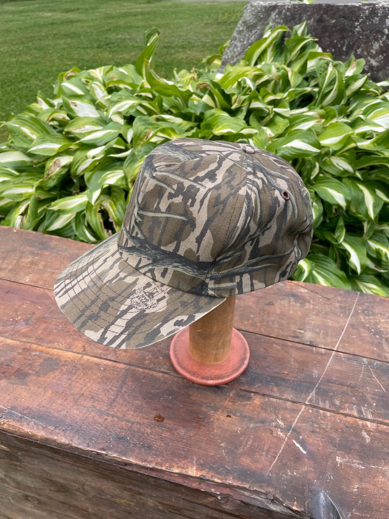 Op de afbeelding: Een baseballpet met een camouflagepatroon in bruin-, beige- en donkergroene tinten. De pet heeft een gestructureerde kroon, een gebogen klep en een klein donker oogje aan de zijkant. De stof is voorzien van een gedetailleerde bosprint.