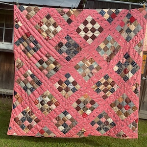 May include: A handmade quilt featuring a pink floral fabric background with diamond-shaped patches of various multi-colored plaid and patterned fabric squares. The quilt is tied with small pink fabric knots at the corners of each square block.