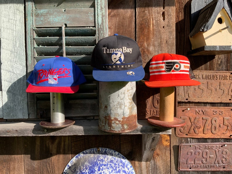 May include: Three vintage baseball caps on display. The left cap is blue with red trim and says "Rangers" in white. The middle cap is black with blue trim and says "Tampa Bay Lightning" in white. The right cap is red with white and black trim and says "Philadelphia Flyers" in white.