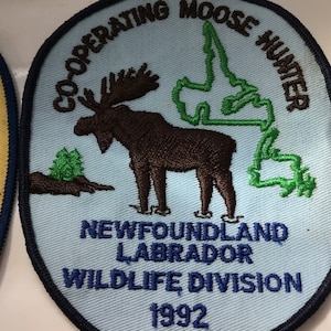 May include: Embroidered patch featuring a brown moose and a green outline of a landmass on a light blue background. Text includes "Co-operating Moose Hunter," "Newfoundland Labrador Wildlife Division," and the year "1992."