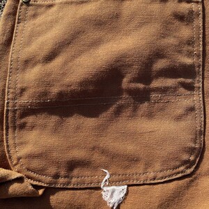 May include: Close-up of a brown canvas pocket on work pants. The pocket is rectangular with a curved bottom and reinforced stitching. A small, white, frayed emblem is sewn near the bottom of the pocket.