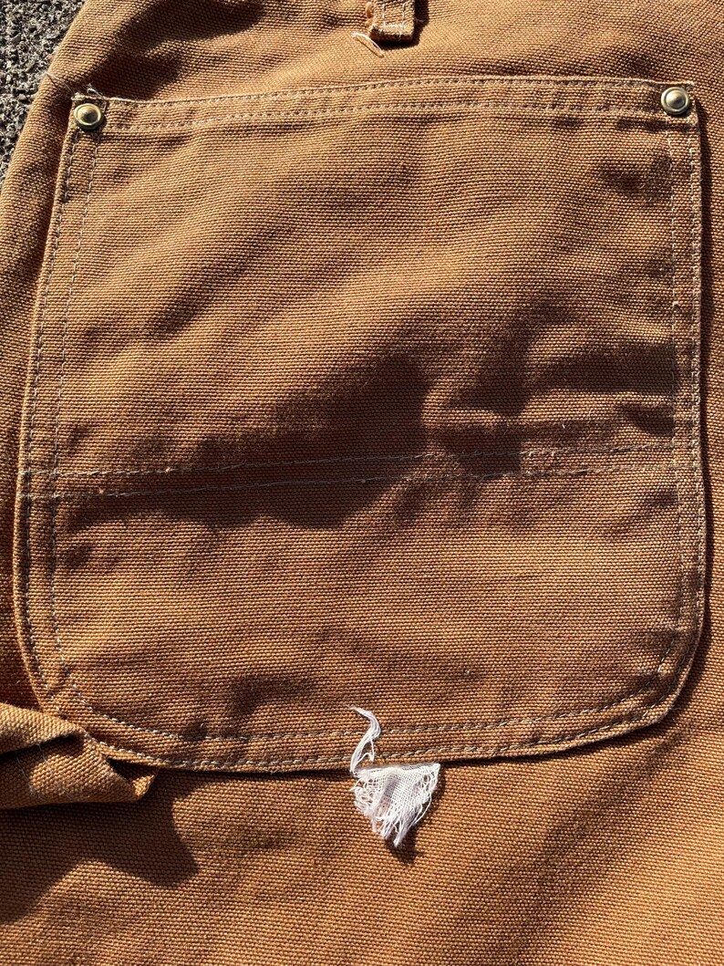 May include: Close-up of a brown canvas pocket on work pants. The pocket is rectangular with a curved bottom and reinforced stitching. A small, white, frayed emblem is sewn near the bottom of the pocket.