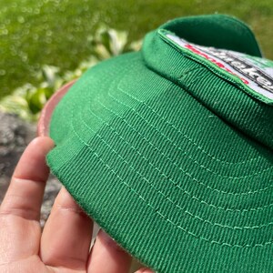 May include: A green baseball cap featuring a white patch with black and red text. The cap is constructed from a textured fabric with visible stitching along the brim and crown, showcasing a structured design with a prominent front panel.