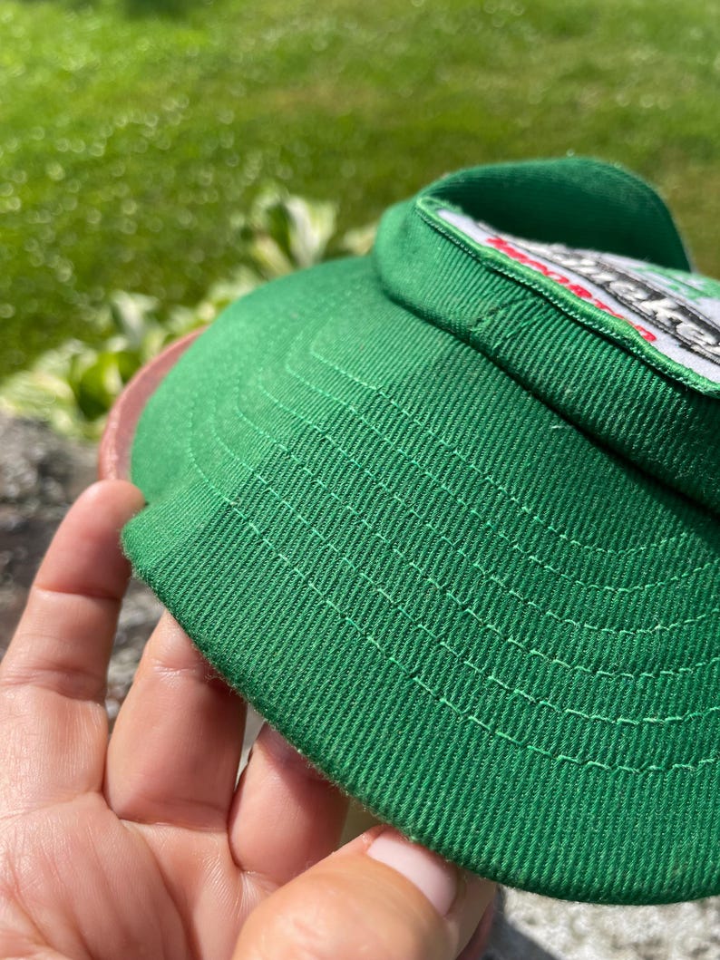 May include: A green baseball cap featuring a white patch with black and red text. The cap is constructed from a textured fabric with visible stitching along the brim and crown, showcasing a structured design with a prominent front panel.