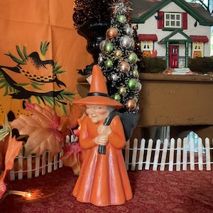 May include: An orange witch figurine with a black sack over her shoulder and a matching hat. The witch is holding a black stick. The figurine is part of a Halloween or fall-themed display, with a small decorative Christmas tree and a miniature house in the background.