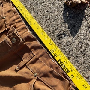 May include: Close-up of brown work pants with a yellow ruler measuring the fabric. The ruler reads "Ellis Fine Home Furnishings" and "159-161 WASHINGTON ST." The pants have visible stitching and metal button details.