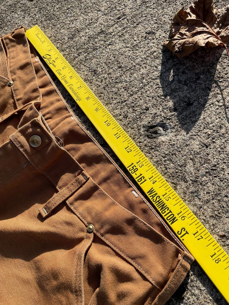 May include: Close-up of brown work pants with a yellow ruler measuring the fabric. The ruler reads "Ellis Fine Home Furnishings" and "159-161 WASHINGTON ST." The pants have visible stitching and metal button details.