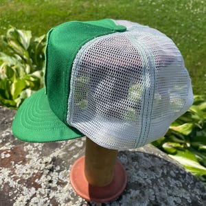 May include: A green and white trucker hat featuring a solid green front panel and brim with a white mesh back panel. The hat has a structured crown and a curved visor with visible stitching detail along the brim and the mesh panels.