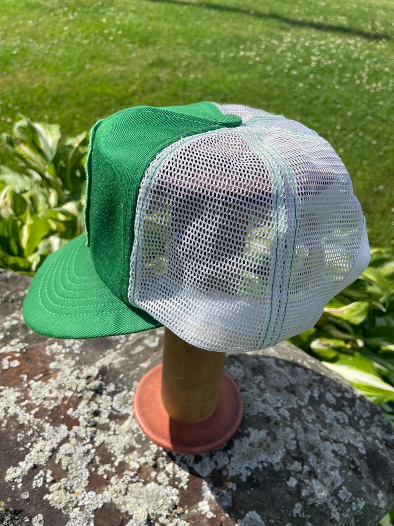 May include: A green and white trucker hat featuring a solid green front panel and brim with a white mesh back panel. The hat has a structured crown and a curved visor with visible stitching detail along the brim and the mesh panels.