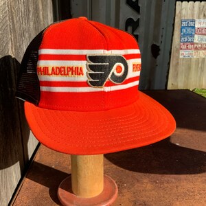 May include: A red, white, and black baseball cap with a mesh back. The front of the cap features a white stripe with the word "PHILADELPHIA" in yellow lettering. A black and orange Flyers logo is centered on the cap.