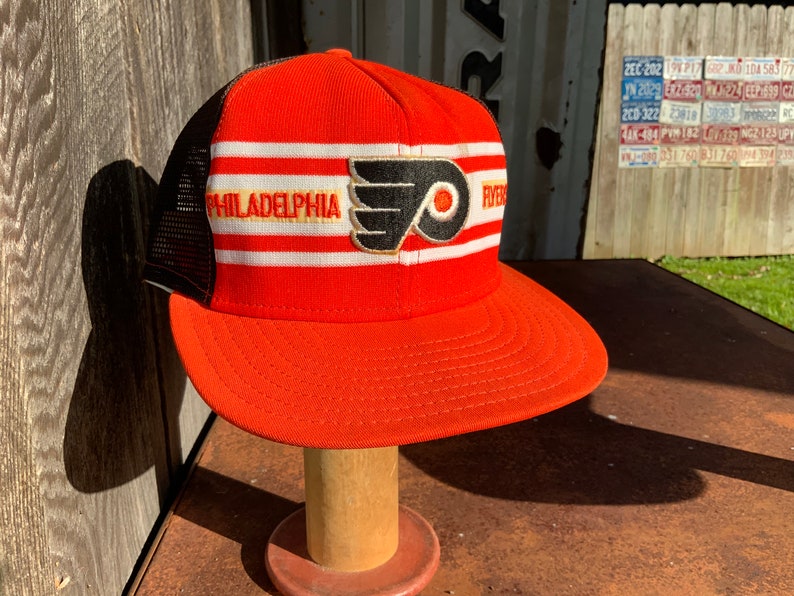 May include: A red, white, and black baseball cap with a mesh back. The front of the cap features a white stripe with the word "PHILADELPHIA" in yellow lettering. A black and orange Flyers logo is centered on the cap.