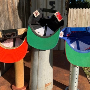 May include: Three baseball caps displayed on stands. The caps are black, red, and blue with green brims. The caps are all turned inside out to show the interior lining.