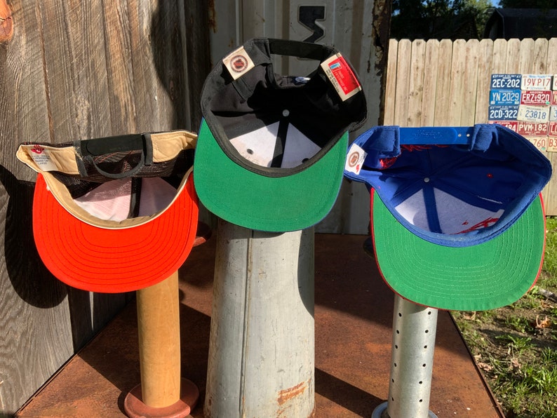 May include: Three baseball caps displayed on stands. The caps are black, red, and blue with green brims. The caps are all turned inside out to show the interior lining.