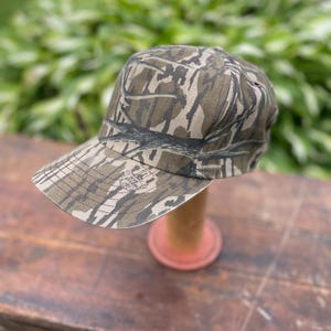 Op de afbeelding: Een baseballpet met een bruin en beige camouflagepatroon met een boomschorsontwerp. De pet heeft een gestructureerde kroon en een gebogen klep. Een klein logo met de tekst Mossy Oak is gedrukt op de linkerkant van de klep bij de kroon.