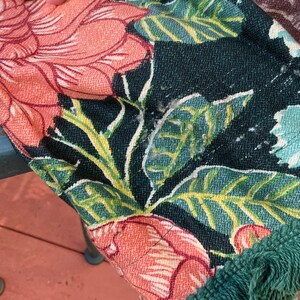 May include: A close-up of a fabric with a floral pattern in shades of pink, green, and white. The fabric has a green fringe border.