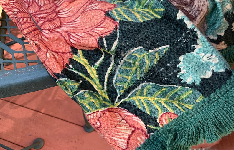 May include: A close-up of a fabric with a floral pattern in shades of pink, green, and white. The fabric has a green fringe border.