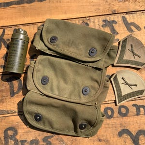 May include: An olive green military canvas pouch with three compartments and black snap closures. A green metal container with text and two olive green patches with crossed rifles are also present. The items are on a wooden surface.