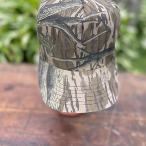 Op de afbeelding: Een baseballpet met een camouflagepatroon van boomschors in bruine, beige en zwarte tinten. De pet heeft een gestructureerde kroon en een gebogen klep met zichtbare stiksels langs de rand. De stof toont een gedetailleerde bosprint.