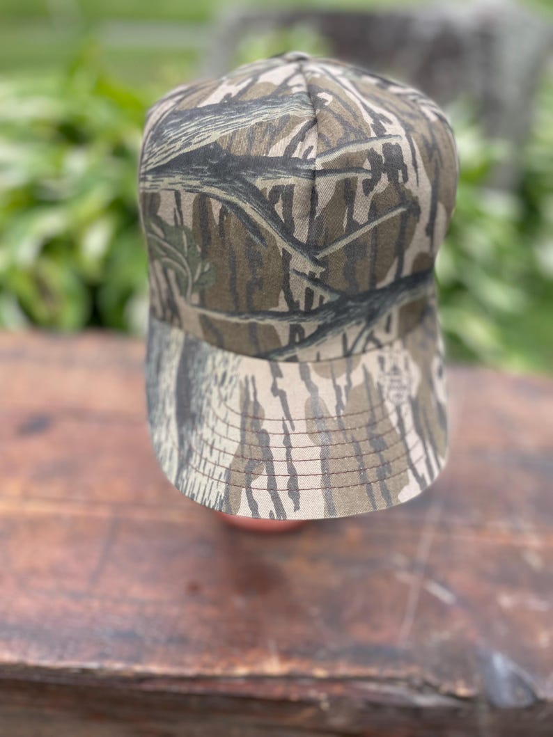 Op de afbeelding: Een baseballpet met een camouflagepatroon van boomschors in bruine, beige en zwarte tinten. De pet heeft een gestructureerde kroon en een gebogen klep met zichtbare stiksels langs de rand. De stof toont een gedetailleerde bosprint.