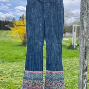 Pu&ograve; includere: Jeans svasati blu denim con orlo a fantasia floreale rosa, verde e blu. I jeans sono appesi a un filo all'aperto contro uno sfondo di cielo nuvoloso ed erba verde.