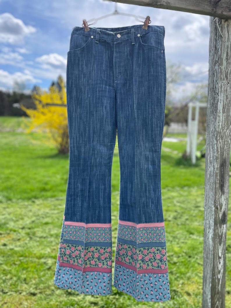Pu&ograve; includere: Jeans svasati blu denim con orlo a fantasia floreale rosa, verde e blu. I jeans sono appesi a un filo all'aperto contro uno sfondo di cielo nuvoloso ed erba verde.