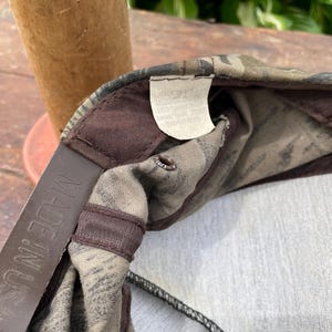 Op de afbeelding: Een baseballpet met camouflageprint en een bruine plastic snapback-sluiting met de tekst MADE IN USA. De binnenkant bevat een wit stoffen label met tekst en bruine stoffen afwerking langs de naden en de ventilatieopeningen.