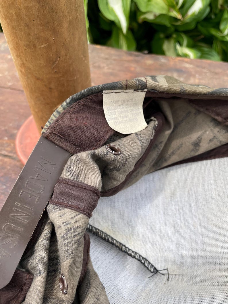Op de afbeelding: Een baseballpet met camouflageprint en een bruine plastic snapback-sluiting met de tekst MADE IN USA. De binnenkant bevat een wit stoffen label met tekst en bruine stoffen afwerking langs de naden en de ventilatieopeningen.