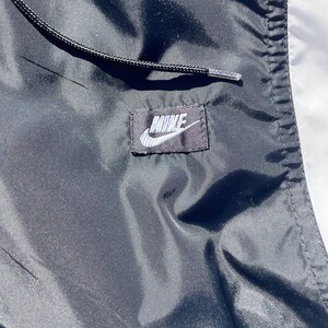 Puede incluir: Primer plano de una chaqueta Nike negra y blanca. La chaqueta presenta un cord&oacute;n negro y un peque&ntilde;o parche rectangular con el logotipo de Nike en blanco. La tela tiene una textura brillante y suave.