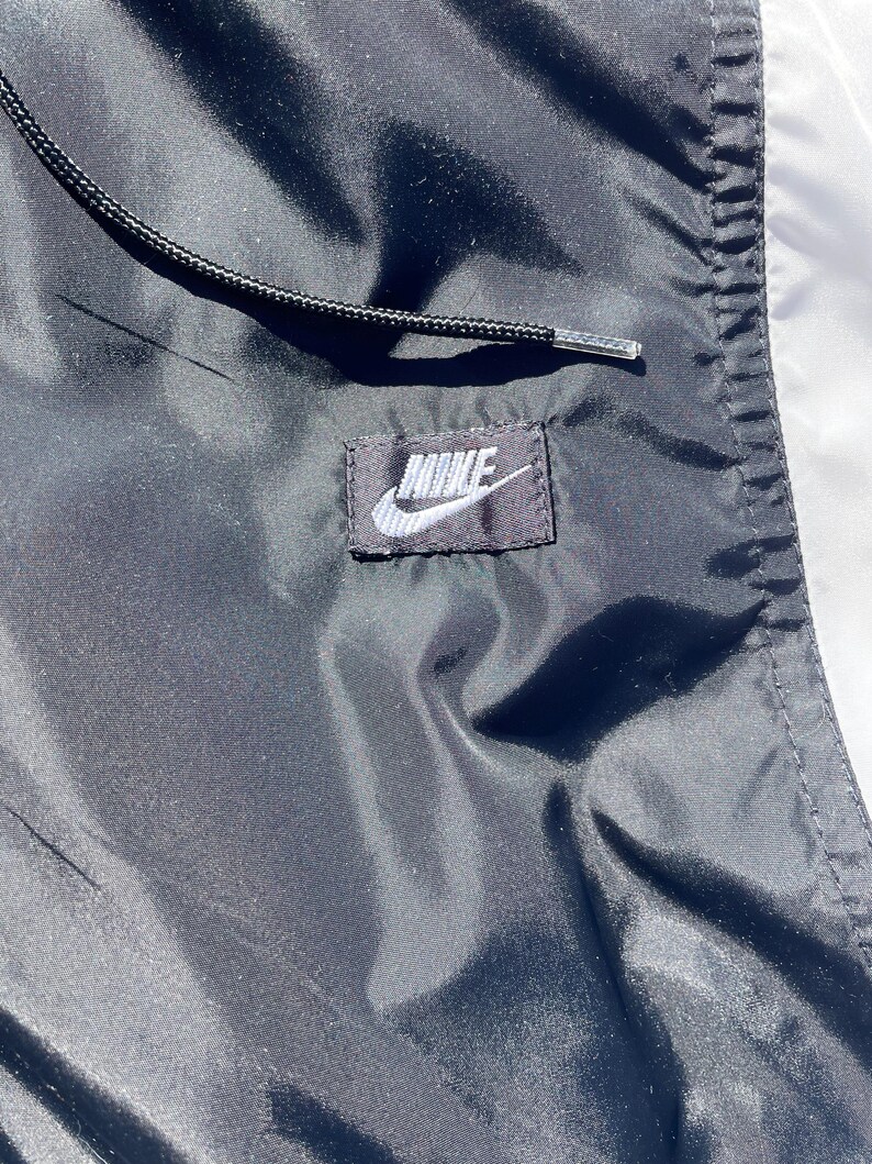 Puede incluir: Primer plano de una chaqueta Nike negra y blanca. La chaqueta presenta un cord&oacute;n negro y un peque&ntilde;o parche rectangular con el logotipo de Nike en blanco. La tela tiene una textura brillante y suave.