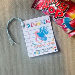 May include: A white paper tag with the words "RING IN A GREAT YEAR!" and "Happy First Day of School!" is shown. The tag features a blue and pink ring pop graphic. A Ring Pop candy wrapper is in the background.