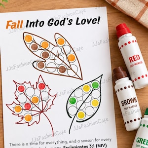 May include: A white activity sheet with the text Fall Into God's Love! and three leaf outlines containing circular spaces for dot markers. Three dot markers in red, brown, and green are positioned to the right of the paper.